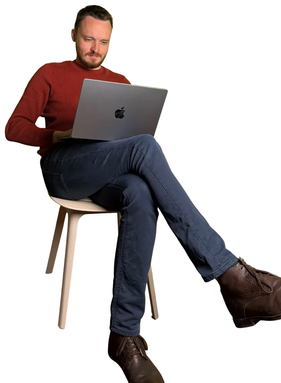Anthony in a red jumper, sitting cross-legged on a chair and working on a laptop